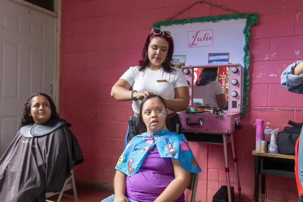 Una estilista peina a una mujer en un salón improvisado con pared rosa, mientras otra espera. Refleja servicios de belleza.