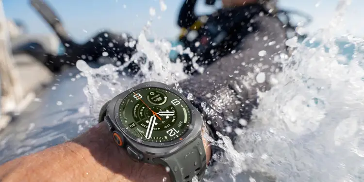 Primer plano de un brazo con un smartwatch sumergido en agua salpicando. En el fondo, una persona con equipo de buceo.