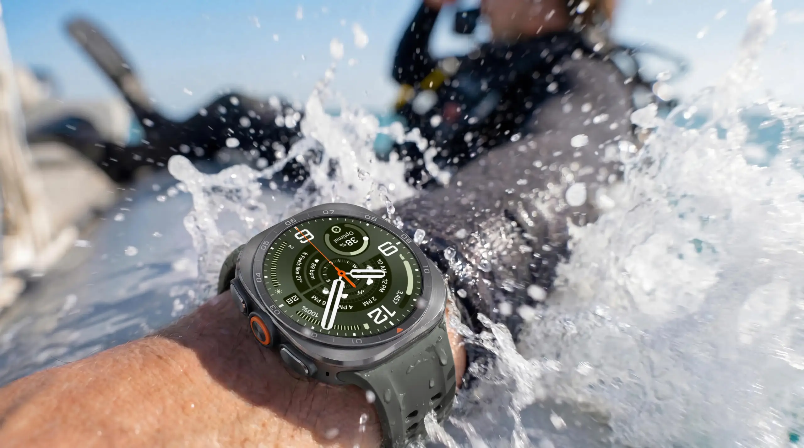 Primer plano de un brazo con un smartwatch sumergido en agua salpicando. En el fondo, una persona con equipo de buceo.