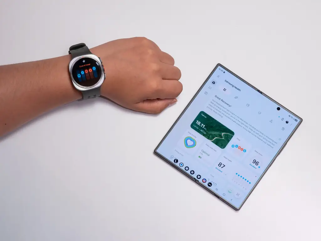 Mano con smartwatch gris mostrando métricas de salud junto a un smartphone plegable con la aplicación Samsung Health abierta, detallando actividad física, recuperación y bienestar.