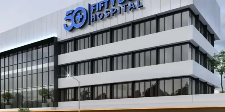 Edificio hospitalario moderno de varios pisos con fachada blanca y grandes ventanales. En la planta baja, locales comerciales y una ambulancia en la calle.