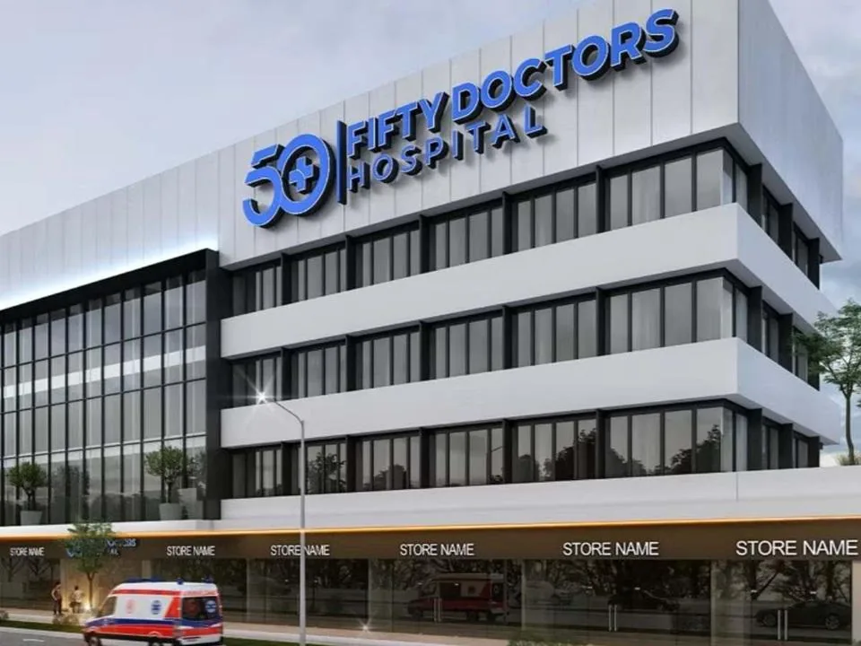 Edificio hospitalario moderno de varios pisos con fachada blanca y grandes ventanales. En la planta baja, locales comerciales y una ambulancia en la calle.