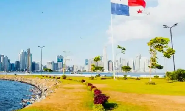 Bandera de Panamá ondeando frente a un moderno skyline de rascacielos junto al mar, con un parque verde en primer plano.