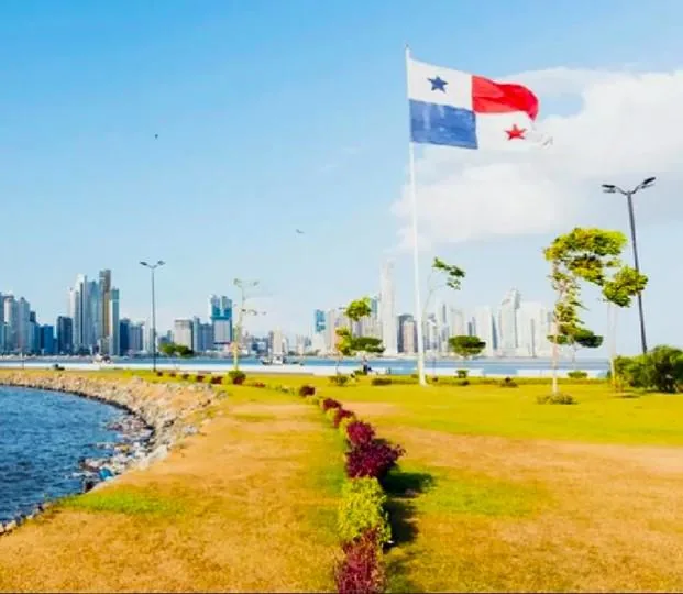 Bandera de Panamá ondeando frente a un moderno skyline de rascacielos junto al mar, con un parque verde en primer plano.