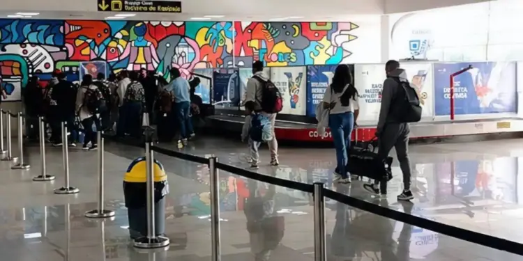 Vista de un área de reclamo de equipaje en un aeropuerto con varias personas esperando sus maletas junto a una cinta transportadora. Un mural colorido adorna la pared.