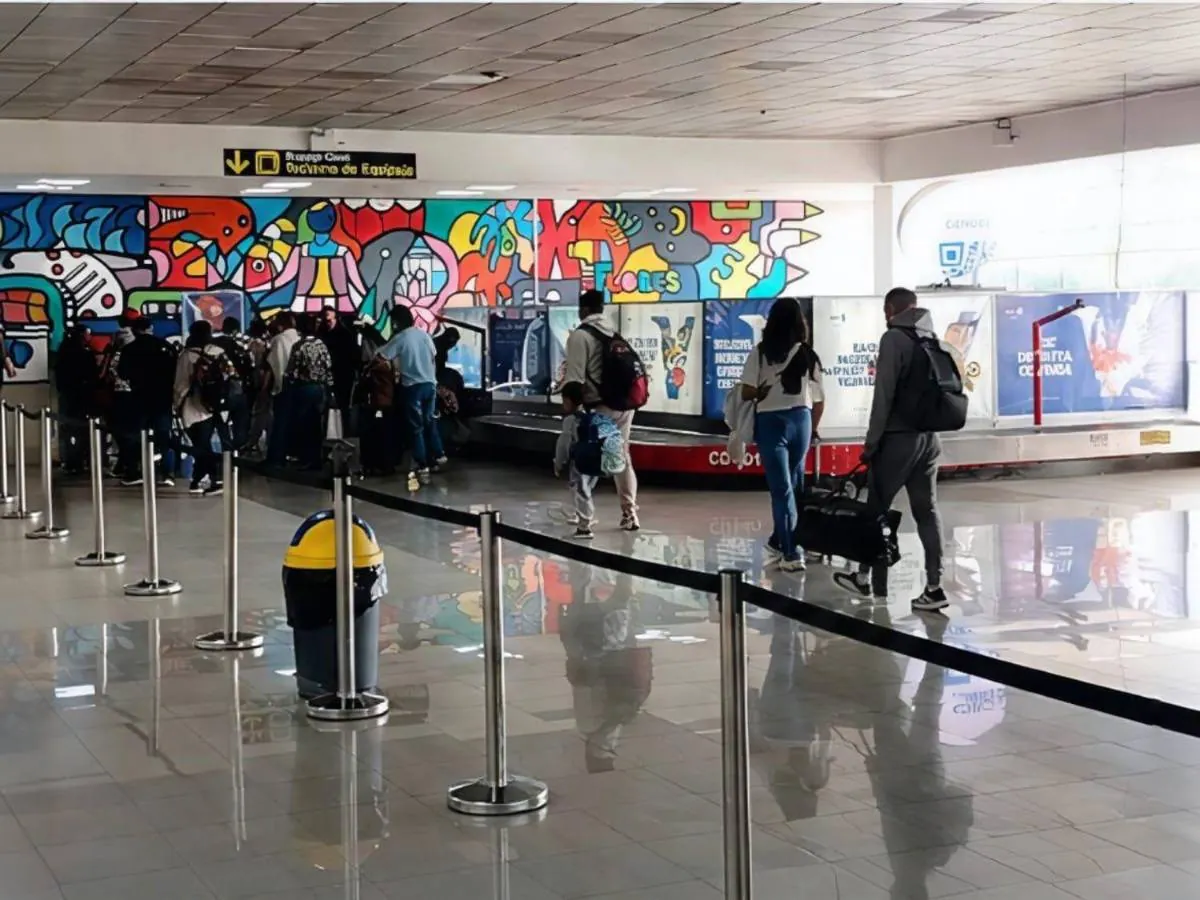 Vista de un área de reclamo de equipaje en un aeropuerto con varias personas esperando sus maletas junto a una cinta transportadora. Un mural colorido adorna la pared.