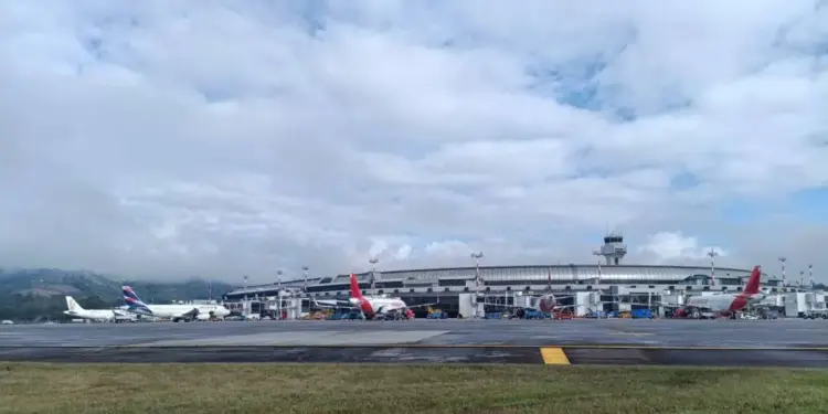 Vista de aeropuerto con aviones LATAM y Avianca en terminal, torre de control, montañas y cielo nublado.