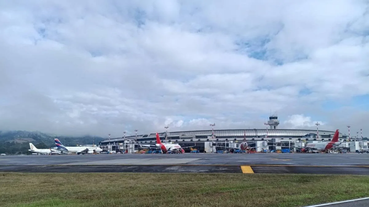 Vista de aeropuerto con aviones LATAM y Avianca en terminal, torre de control, montañas y cielo nublado.