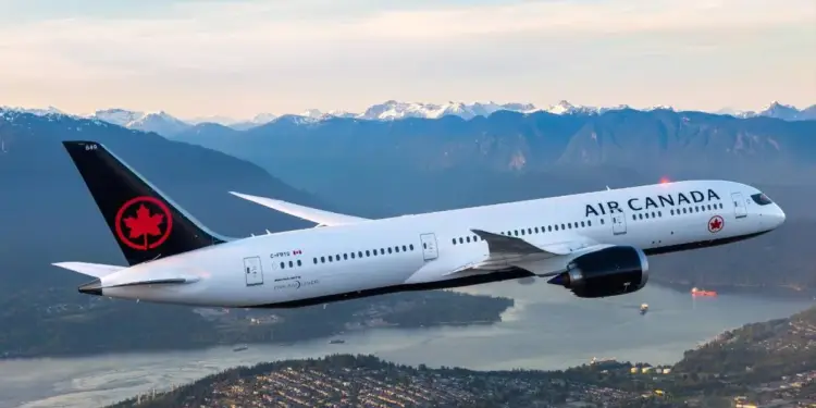 Avión blanco de Air Canada volando sobre una ciudad costera con montañas nevadas al fondo. Refleja transporte aéreo y logística.