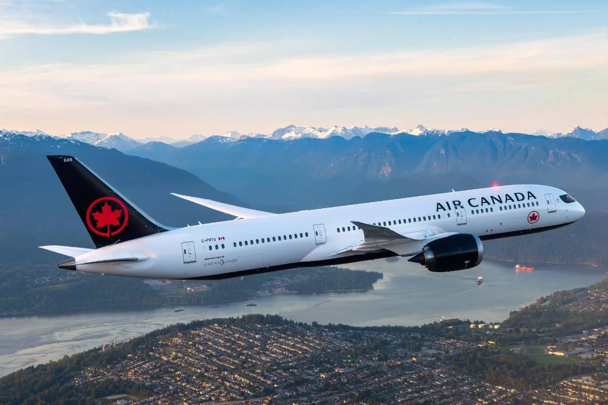 Avión blanco de Air Canada volando sobre una ciudad costera con montañas nevadas al fondo. Refleja transporte aéreo y logística.