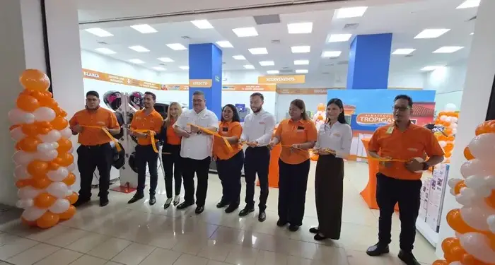 Grupo de ocho personas, incluyendo hombres y mujeres, cortando una cinta naranja en la inauguración de una tienda. Hay globos naranjas y blancos. Se ve un letrero de 'Almacenes Tropigas'.