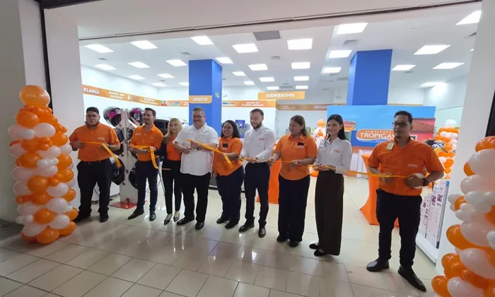 Grupo de ocho personas, incluyendo hombres y mujeres, cortando una cinta naranja en la inauguración de una tienda. Hay globos naranjas y blancos. Se ve un letrero de 'Almacenes Tropigas'.