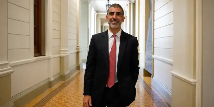 Un hombre de negocios con traje negro, camisa blanca y corbata roja sonríe mientras camina por un pasillo con arquitectura clásica y suelo decorado.
