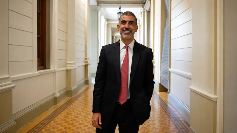 Un hombre de negocios con traje negro, camisa blanca y corbata roja sonríe mientras camina por un pasillo con arquitectura clásica y suelo decorado.