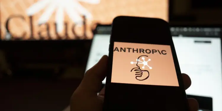 Una mano sostiene un smartphone mostrando el logo de Anthropic con su nombre y un icono de cabeza conectada. Al fondo, una pantalla borrosa con el texto 'Claud'.
