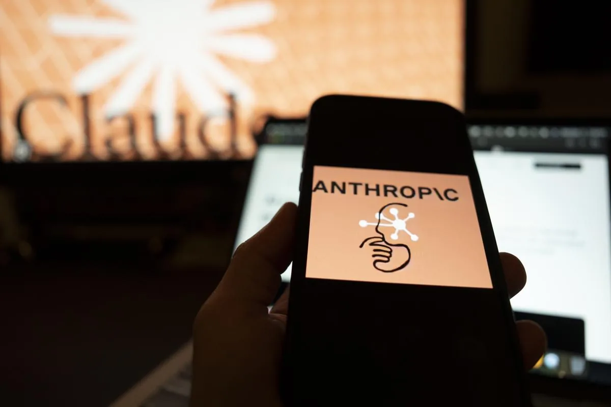 Una mano sostiene un smartphone mostrando el logo de Anthropic con su nombre y un icono de cabeza conectada. Al fondo, una pantalla borrosa con el texto 'Claud'.