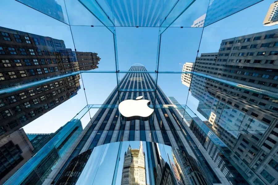 Vista en contrapicado de un cubo de cristal transparente con un logotipo corporativo en el centro, rodeado de rascacielos y cielo azul.