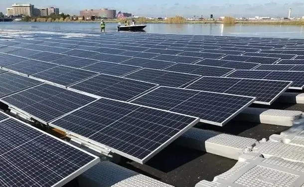 Amplia instalación de paneles solares flotantes sobre el agua, con estructuras de soporte. Un bote y edificios en el fondo.