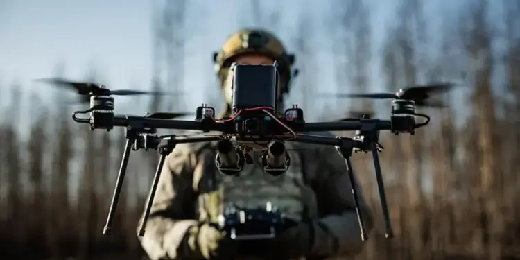 Primer plano de un dron profesional con múltiples rotores, sostenido por una persona con equipo táctico que opera un control remoto. Fondo borroso de árboles.