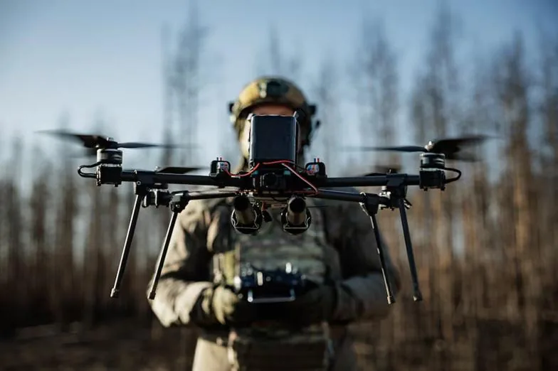 Primer plano de un dron profesional con múltiples rotores, sostenido por una persona con equipo táctico que opera un control remoto. Fondo borroso de árboles.