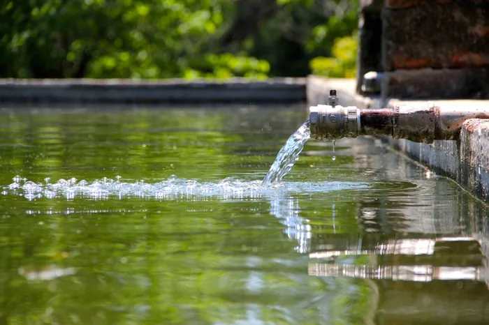 Agua fluyendo desde una tubería metálica hacia un estanque exterior, representando el suministro y uso de recursos hídricos.