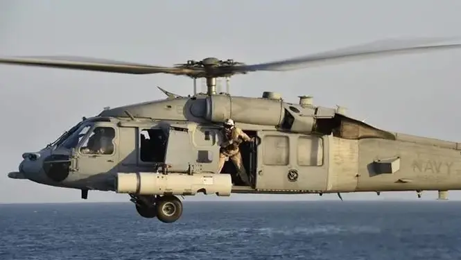 Un helicóptero militar gris vuela sobre el mar con un tripulante en la puerta abierta.