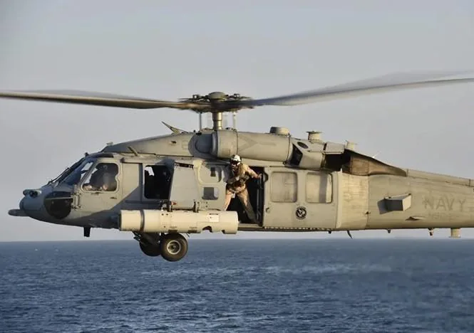 Un helicóptero militar gris vuela sobre el mar con un tripulante en la puerta abierta.