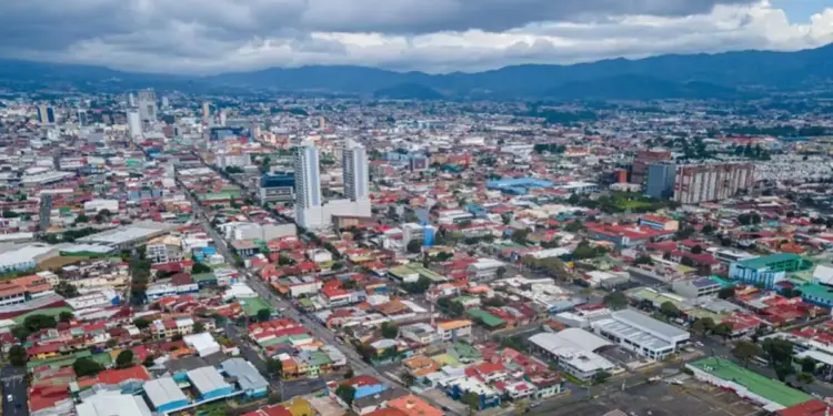Vista aérea de una ciudad densamente poblada con edificios residenciales y comerciales, calles transitadas y montañas al fondo bajo un cielo nublado. Refleja crecimiento urbano.