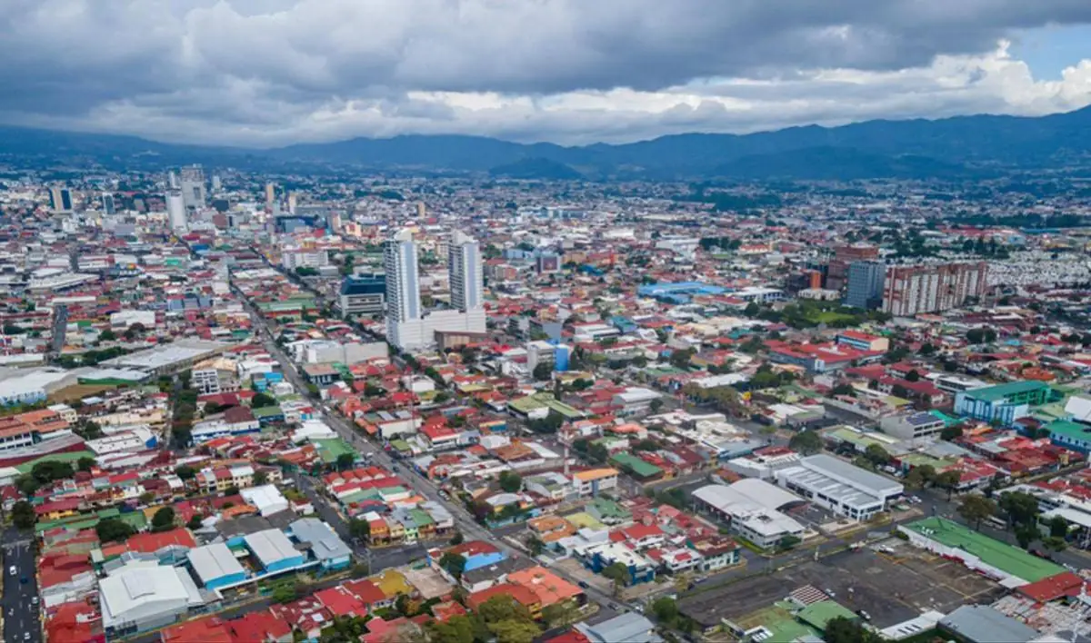 Vista aérea de una ciudad densamente poblada con edificios residenciales y comerciales, calles transitadas y montañas al fondo bajo un cielo nublado. Refleja crecimiento urbano.