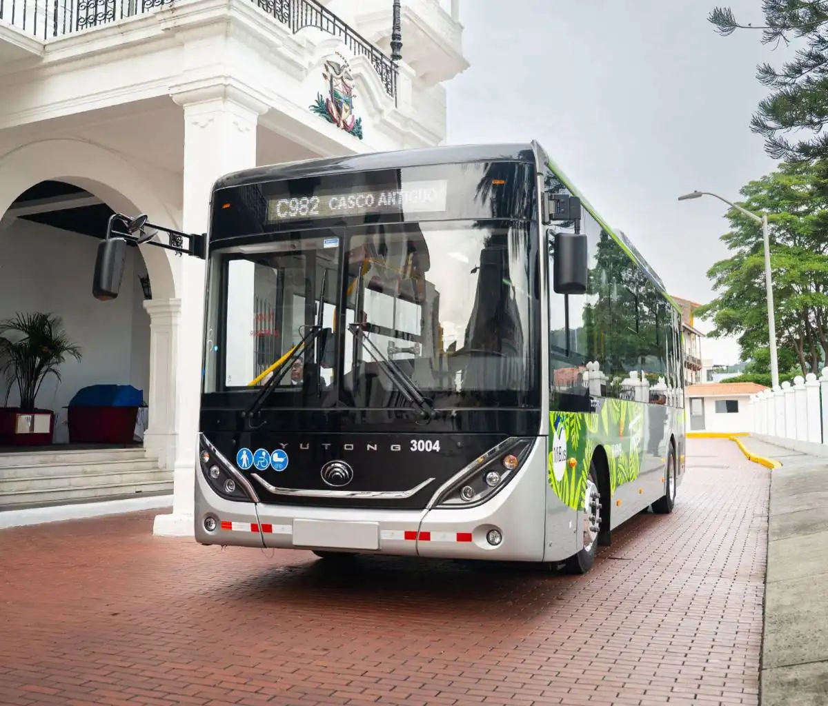 Autobús de transporte público moderno detenido en una calle adoquinada frente a un edificio con columnas blancas.