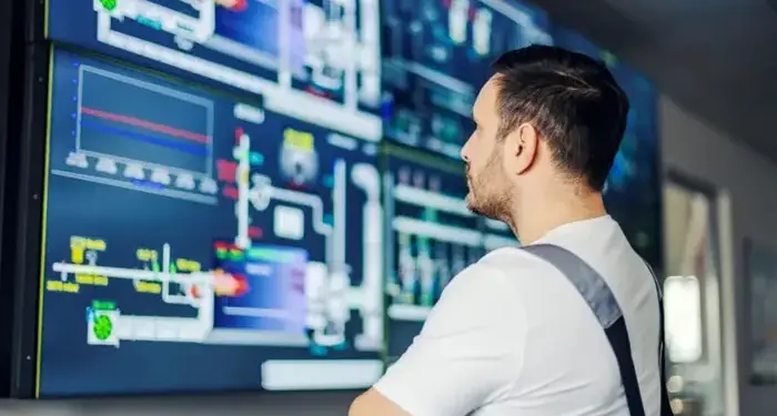 Trabajador observando grandes pantallas con diagramas de flujo y gráficos analíticos en una sala de control.