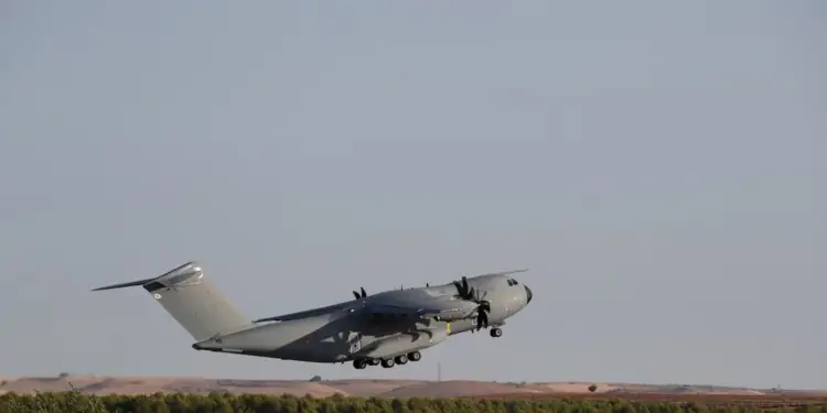 Avión de transporte militar de color gris despegando sobre una zona boscosa bajo un cielo claro.