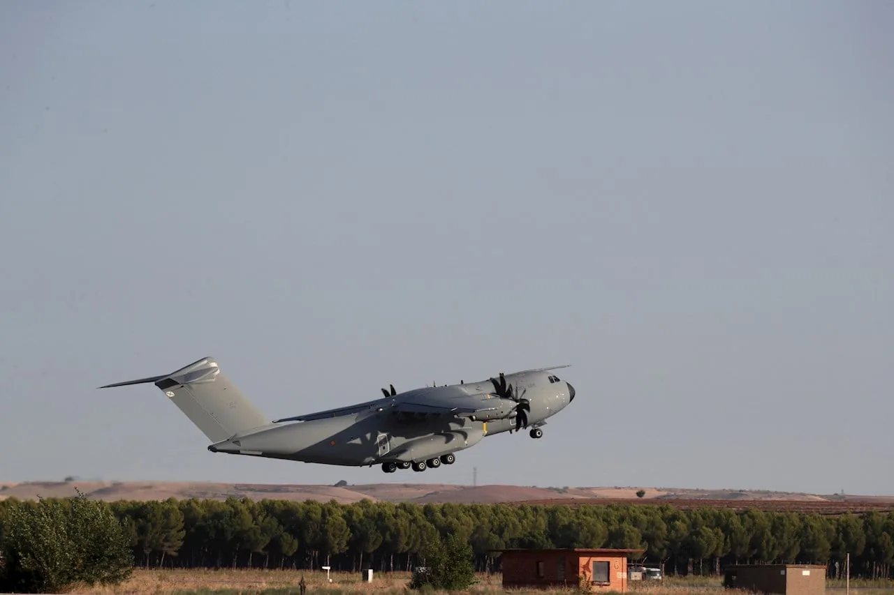 Avión de transporte militar de color gris despegando sobre una zona boscosa bajo un cielo claro.