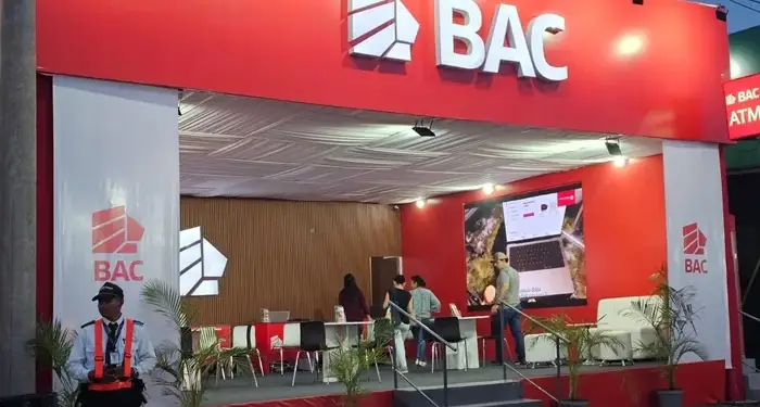 Vista frontal de un stand moderno de BAC en rojo y blanco con su logo, un guardia de seguridad y personas interactuando dentro. Incluye un ATM.