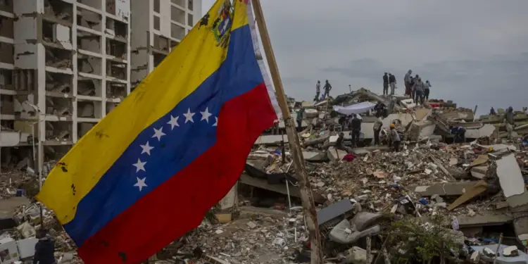 Bandera de Venezuela ondeando frente a un edificio dañado y escombros de una estructura colapsada con personas en el fondo