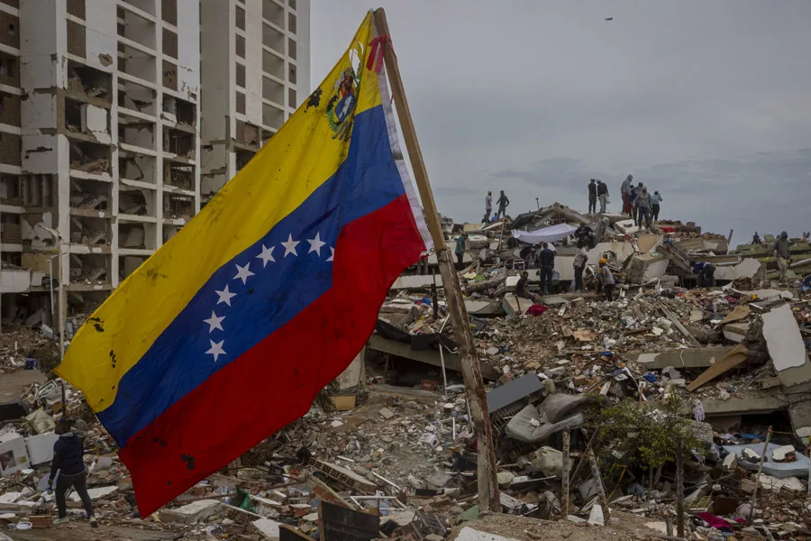 Bandera de Venezuela ondeando frente a un edificio dañado y escombros de una estructura colapsada con personas en el fondo