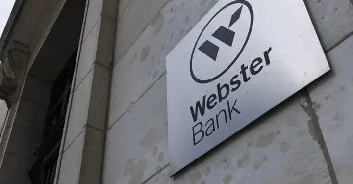 Primer plano del letrero metálico de Webster Bank en la fachada de un edificio de piedra clara. Se ve el logo y el nombre del banco.