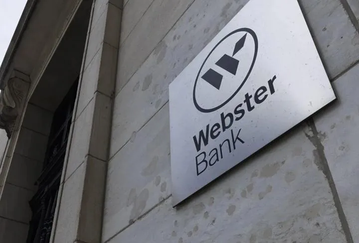 Primer plano del letrero metálico de Webster Bank en la fachada de un edificio de piedra clara. Se ve el logo y el nombre del banco.