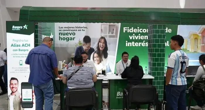 Personas interactuando en un stand de Banpro con banners sobre registro de Alias ACH y fideicomisos de vivienda de interés social.