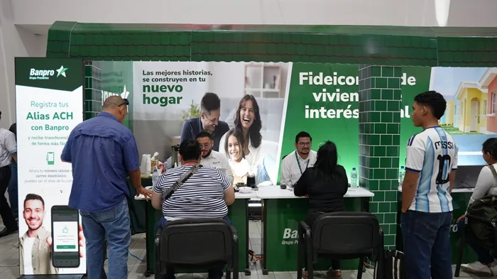 Personas interactuando en un stand de Banpro con banners sobre registro de Alias ACH y fideicomisos de vivienda de interés social.