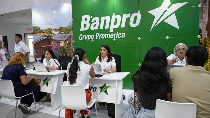 Personal de Banpro Grupo Promerica asesora a clientes en un stand con laptops, frente a un gran logo verde y casas con paneles solares.