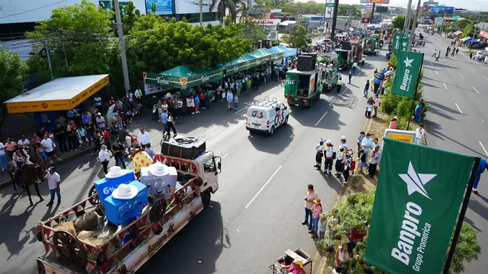 Vista aérea de un desfile urbano con multitudes a los lados de la calle. Se observan camiones, una van y banderas verdes de Banpro Grupo Promerica.