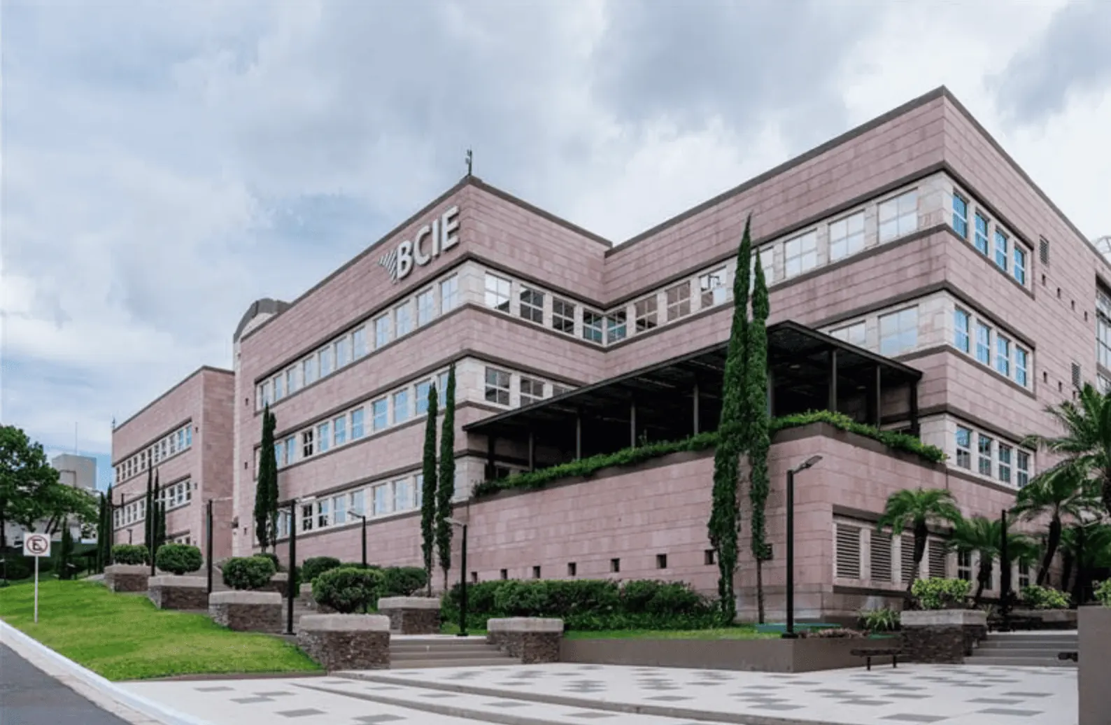 Vista exterior de un moderno edificio de oficinas con fachada de piedra rosada y múltiples ventanas. El letrero 'BCIE' es visible en la parte superior. Rodeado de vegetación, árboles y un camino pavimentado.