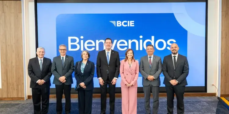 Grupo de siete ejecutivos, cuatro hombres y tres mujeres, posando frente a una pantalla azul con el logo BCIE y la palabra 'Bienvenidos'. Vestimenta formal de negocios.