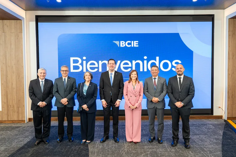 Grupo de siete ejecutivos, cuatro hombres y tres mujeres, posando frente a una pantalla azul con el logo BCIE y la palabra 'Bienvenidos'. Vestimenta formal de negocios.
