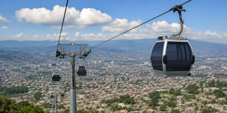 Vista aérea de un teleférico moderno con varias cabinas sobre una extensa área urbana con casas y edificios, montañas al fondo.
