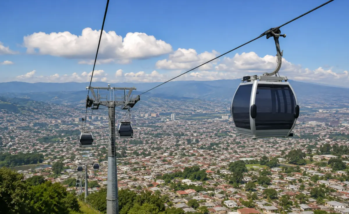 Vista aérea de un teleférico moderno con varias cabinas sobre una extensa área urbana con casas y edificios, montañas al fondo.