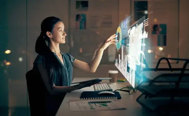 Mujer profesional interactuando con una pantalla holográfica de datos financieros y gráficos en una oficina moderna. Sostiene una tablet y hay un teclado y ratón en el escritorio.