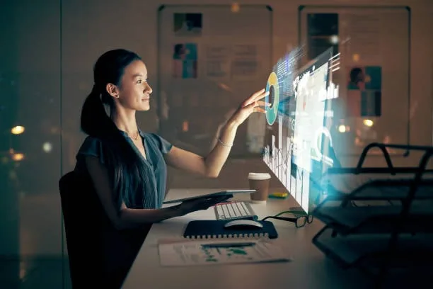 Mujer profesional interactuando con una pantalla holográfica de datos financieros y gráficos en una oficina moderna. Sostiene una tablet y hay un teclado y ratón en el escritorio.