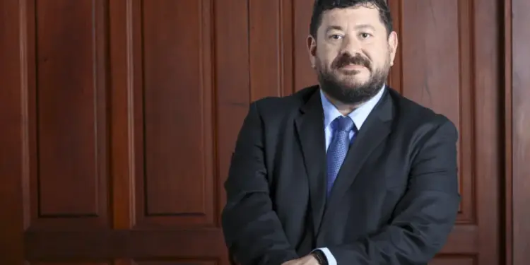 Hombre profesional con barba y bigote, vestido con traje oscuro y corbata azul, posando con las manos sobre una silla frente a una pared de madera.